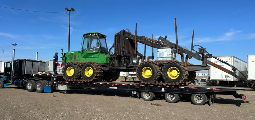 Green John Deere timber forwarder loaded on a flatbed semi-truck trailer for heavy equipment shipping managed by trained operators.